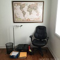 Photo of National Geographic world map mounted above black office chair, Canon printer, floor lamp, waste bin, envelopes, and office supplies arranged near the chair.
