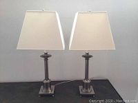 Two matching metal table lamps with white square shades standing side by side on a dark surface.