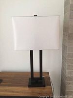 Front view of the Clarke iron base table lamp with white rectangular shade on a wooden surface against a light wall.