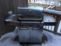 Front view of grill with hood closed showing control panel, doors and side shelves in snowy outdoor setting