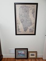 Framed New York City map hung on wall with two smaller framed artworks resting below on the floor.