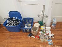 Mixed household items spread on floor including a blue VAVA VA AH012 humidifier base (no remote), ceramic pot, watering can, metal shower shelf, various tapes, electrical cords, and small hardware supplies placed in blue plastic bins and baskets.