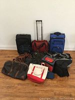 Full lot of eleven travel bags including several rolling suitcases and duffle bags arranged against a white wall on a wooden floor.