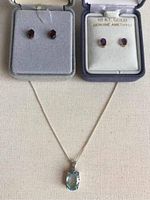 Two pairs of stud earrings in velvet boxes with a silver necklace with blue pendant below on a cloth background.