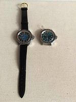 Photo of two Vostok Komandirskie men's watches, one on black leather strap, one without a band, laid flat on a cream fabric surface.