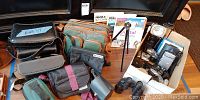 Wide photo showing multiple camera bags, cameras, flashes, binoculars, photography books, tripods, and accessories on a table.