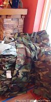 Photo of folded stack and one unfolded pair of woodland camouflage chemical protective pants with suspenders.