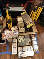 Photo showing lot contents including four yellow tripods, various boxed electrical ballasts and power supplies, packages of connectors, wiremold boxes, and boxed tools.