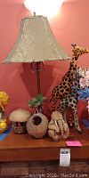Floor lamp with beige fabric shade and multiple figurines including giraffes and coconut shell art on a wooden surface against a pink wall