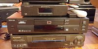 Photo showing four electronic devices stacked: Natvision digital satellite receiver on top, Toshiba DVD player in middle, Panasonic VHS player at bottom, with remotes on top right.