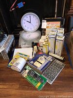 Photo showing white-faced wall clock on bubble wrap, various boxed light bulbs, universal remotes, home security sensors, a calculator, and flashlights on a wooden surface.