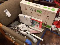 Photo showing the Wii Fit balance board box, Wii controllers, sensor bar, and stacked game cases on a chair.