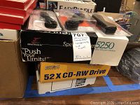 Photo showing SpeedStream 5250 modem box, three black cables in plastic packaging, and stacked boxed networking items including a 52X CD-RW Drive.