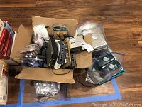 Box containing various electronic items including DVD drives, USB cables, switches, routers, and PC memory cards scattered inside and partially visible.