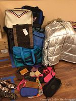 Full view of silver Victorious Homme puffer coat, turquoise duffle, stacked travel bags, and assorted small bags and wallets on floor
