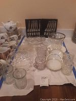 Photo showing multiple glass items including plates, knives in case, bowls, and mugs arranged on a table.