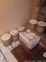 Multiple stacks of white fine china plates with gold trim next to a cardboard box full of small cup pieces on a wooden surface.