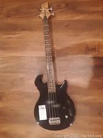 Full body view of black Wildfire Drive electric bass guitar on wooden floor, showing four strings, bridge, knobs, and overall body shape with chrome hardware.