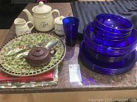 Assorted kitchenware including blue glass dishes stacked, ceramic tea set, mosaic serve plate, utensils, and blue glass cup on countertop.