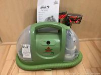 Front view of the Bissell Little Green Machine carpet cleaner showing green body, clear plastic water tank, attached hose coiled at base and cleaning nozzle with red bristles. User manual also visible behind the unit.