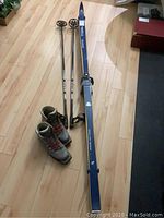 Full set of cross country skiing equipment laid out on wooden floor: one pair of skis with bindings, two poles with black baskets, and a pair of gray ski boots.