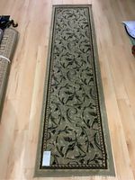 Full length photo showing green carpet runner with floral vine pattern and border on light wood floor.