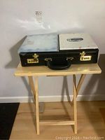 Black briefcase with combination locks and two designer stationery boxes resting on top of a folding wooden TV table.