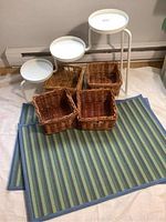 Triple plant stand with three round white trays, four wicker baskets, and two striped area mats arranged in front.