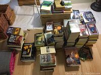 Wide angle view showing multiple stacks of hardcover and paperback books including science fiction and fantasy titles, with a tote bin on top of a cardboard box.