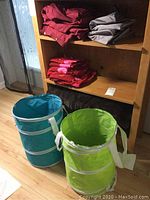 Two collapsible laundry baskets, one blue and one green, on a floor in front of wooden shelving filled with bedding linens in red, burgundy, gray and patterns.