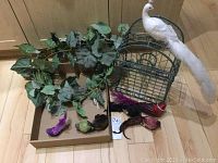 Green wire bird cage with white and colorful decorative birds and artificial grape leaf vine on wood floor.