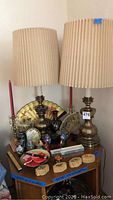 Photo of two tall brass lamps with large pleated beige lampshades. Multiple decorative oriental-style fans and assorted jars and ceramic items on a wooden surface underneath.