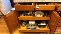 Wooden kitchen cabinet with open drawers and doors showing various kitchenware items including bakeware and flatware.