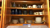 Wooden cabinet shelf showing stacked plates, black bowls, blue ceramic mugs, printed white mugs and glassware