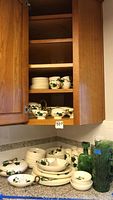 Overall view of Metlox dish set stored in a wooden cabinet showing stacked plates, bowls, and cups with green vine design.
