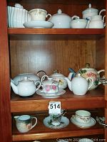 Wooden cabinet shelves holding assorted teapots, teacups, and saucers in white and floral patterns of varying styles.