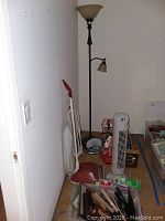 Floor lamp with dual light fixtures next to Hoover vacuum and various other items including tower fan, batteries, heat lamp, light bulbs, clock, and picture frames on the floor.
