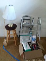 Photo showing two metal walkers, wooden stool, and two lamps with white shades; box of assorted bathroom and medical supplies in front