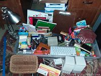 Photo showing a clear plastic container filled with assorted office supplies, an adjustable silver metal desk lamp, a wired keyboard, and a small basket in the foreground.