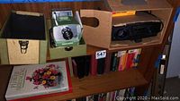 Shelf displaying two vintage projectors, a projection screen, and vintage books below