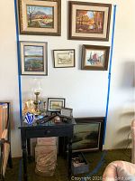 Overall view of black wooden table with glass top under six framed paintings and framed pictures on wall and floor, also showing assorted decorative and functional items on and around the table.