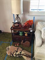Photo showing a small wooden side table with various art objects and a mid-century style table lamp with a white shade on top. Also visible is a large orange translucent fish-shaped sculpture or bowl, and ceramic bowls on the shelf below.