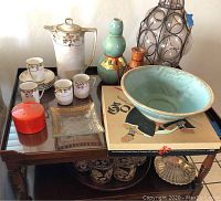 Photo showing a wooden table with various items including a white porcelain coffee pot and cups, turquoise ceramic bowl, decorative ceramic vases, vintage Go board game, glass dishes, and a large clear glass bottle with metal rings.