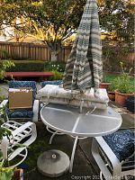 Wide view of the patio set showing table with glass top, one metal chair with blue paisley cushion, umbrella closed and standing in center of table, extra cushions and box with slip covers on table.