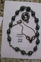 Photo of large oval stone bead necklace with blue, green, and black hues, arranged in oval shape with clasp and metal chain extension visible on white background.