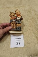 Front view of the Goebel Hummel Brothers figurine held in hand on beige cloth background showing the two boys with arm around each other.