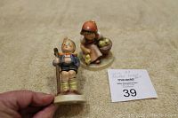 Two German Goebel Hummel figurines placed on a beige textured surface, showing size and condition with a MaxSold tag.