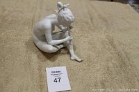 Full profile view of white bisque porcelain figurine of a seated girl playing flute on beige fabric background.