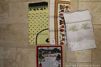 Photo showing all four textile pieces folded and laid on the floor: green chicken table cloth, orange multicolored table cloth, white pine branch table cloth, and Bavarian tea towel