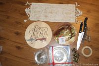 Overview of lot items including wooden cheese board, decorative plate, knives, kitchen utensils, stainless pot, table runner.